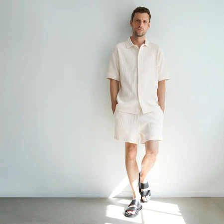 Man in matching cream-colored shirt and shorts stands against a white wall in a casual pose wearing black sandals. Minimalist fashion.