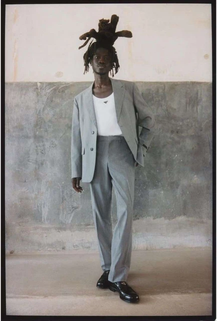 An adult male with dreadlocks wearing a grey suit and black shoes stands in a sparsely furnished room with a worn wall.