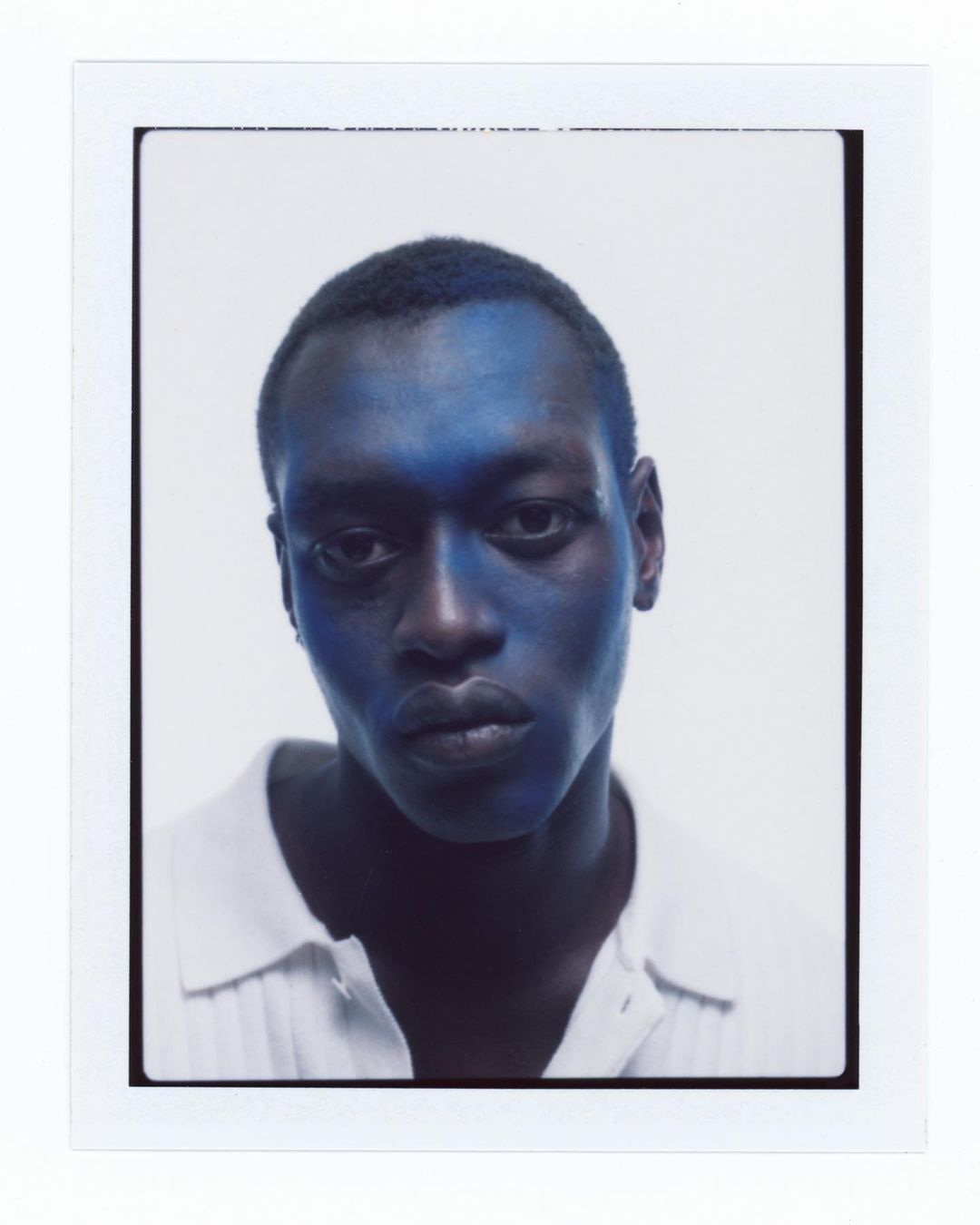 Portrait of a Black male model with blue face paint, wearing a white collared shirt against a bright white studio backdrop.