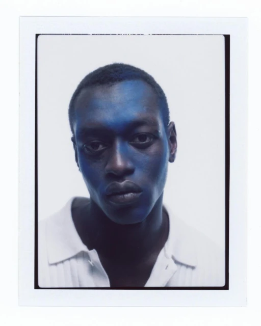 Portrait of a Black male model with blue face paint, wearing a white collared shirt against a bright white studio backdrop.