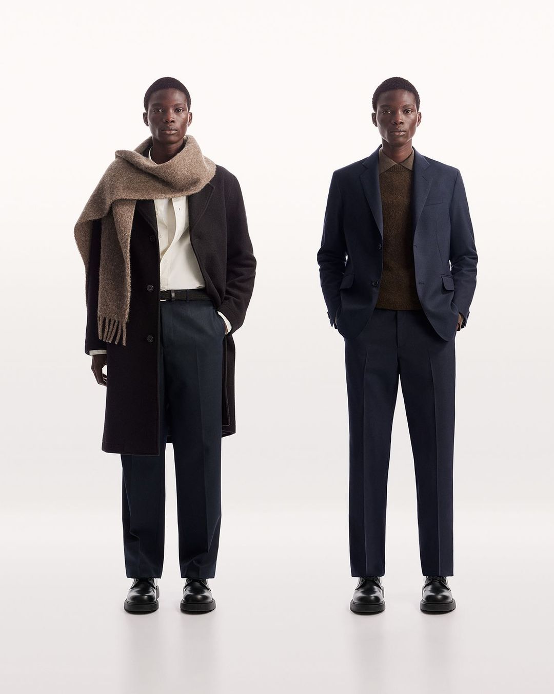 Two adult Black men in formal attire, one with a wool scarf, stand in a minimalist studio against a bright white backdrop, showcasing fashion.