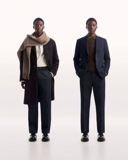 Two adult Black men in formal attire, one with a wool scarf, stand in a minimalist studio against a bright white backdrop, showcasing fashion.
