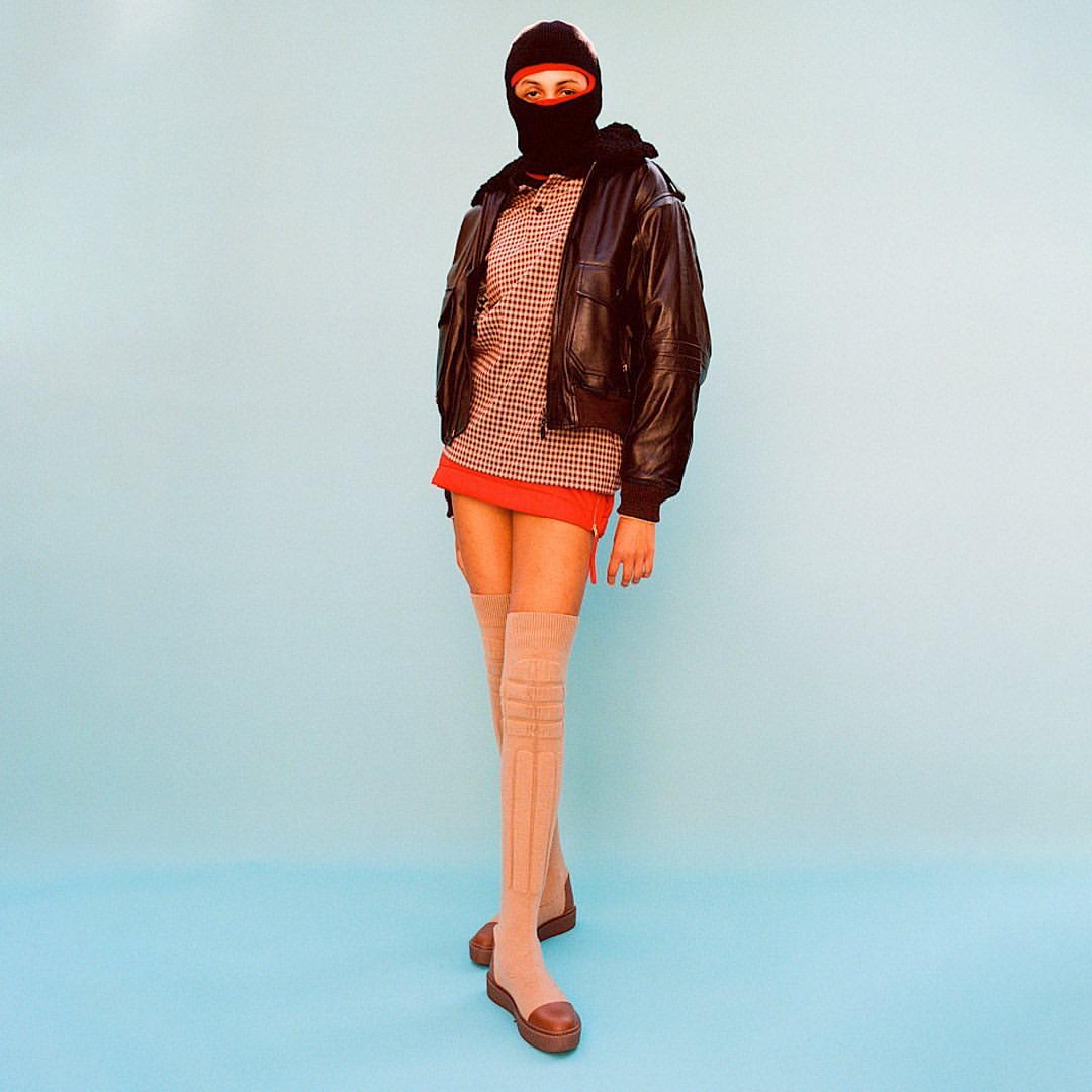 Full-length portrait of a person in a black balaclava, jacket, mini skirt, and thigh-high socks against a blue background, studio lighting.