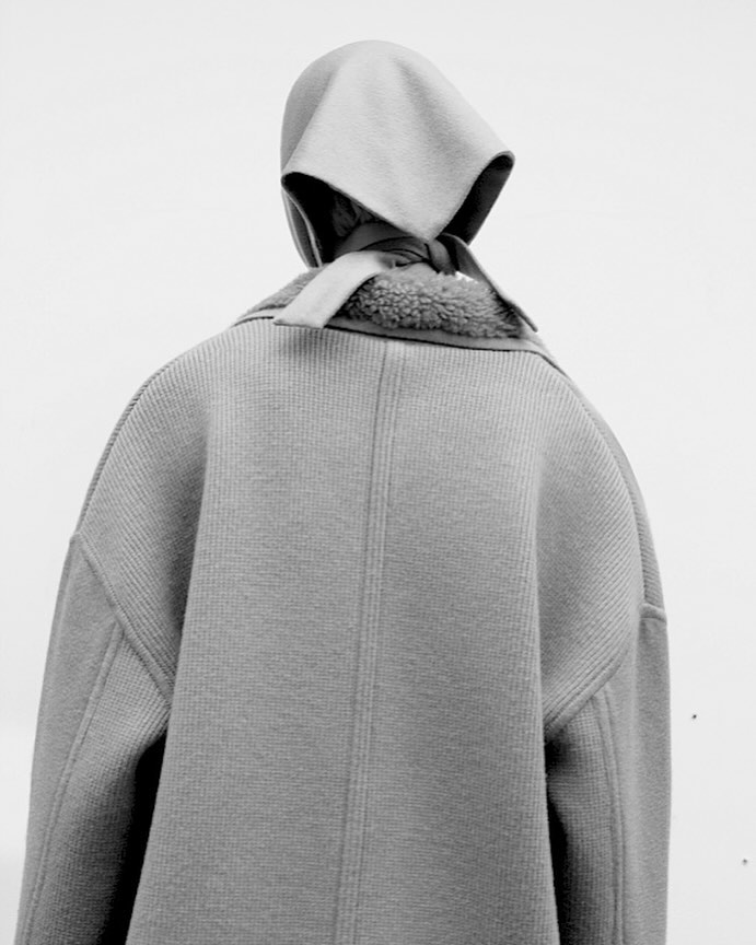 Person from the back wearing a neutral-toned coat with a sherpa collar and a matching bonnet-style hat against a white backdrop.
