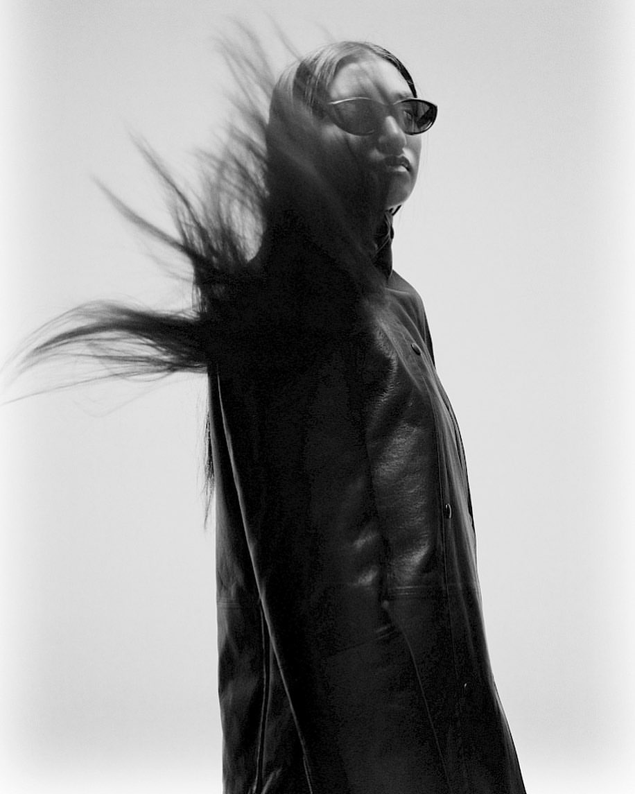 Black and white portrait of a woman with long hair blowing in the wind, wearing sunglasses and a leather jacket against a bright background.