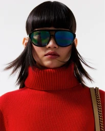 Model wearing dark mirrored aviator sunglasses and a bright red turtleneck sweater against a clean white backdrop.