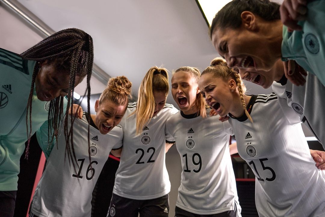 German women's soccer team huddle together in team jerseys, shouting with arms around each other in a tunnel or locker room setting.