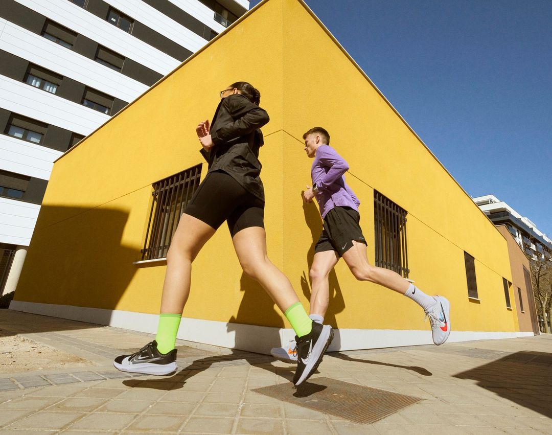 Two athletic adults in running attire are actively jogging on a paved surface with a yellow building and modern architecture in the backdrop.