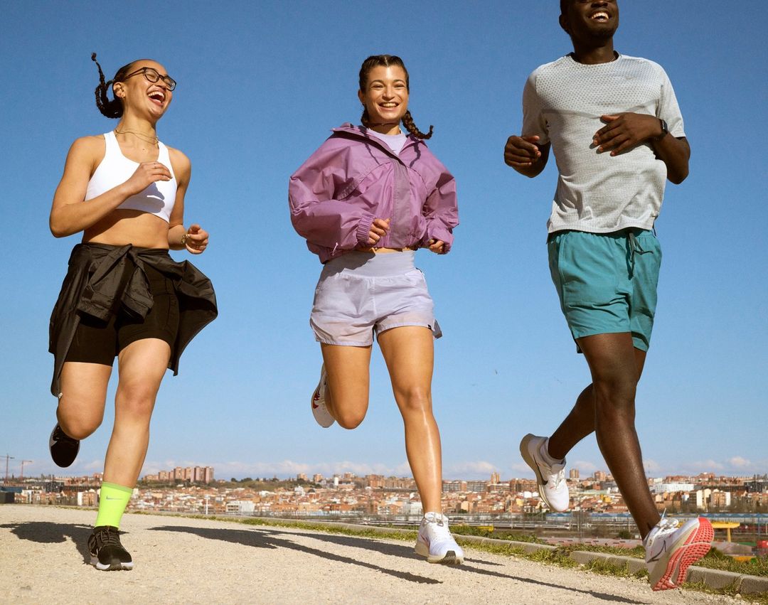 Three diverse adults joyfully running outdoors on a sunny day, dressed in athletic wear, with a blurred cityscape in the background.