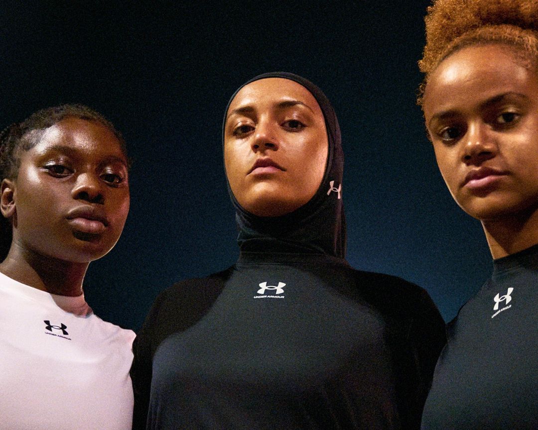 Three athletic women of diverse ethnicities in sports apparel face the camera in a close-up shot against a dark background. All wear Under Armour branded shirts.