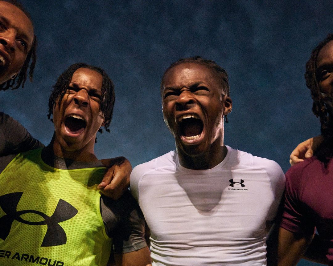 Four Black male athletes, tightly framed, intensely shout with visible Under Armour shirts, suggesting a moment of competition or celebration.