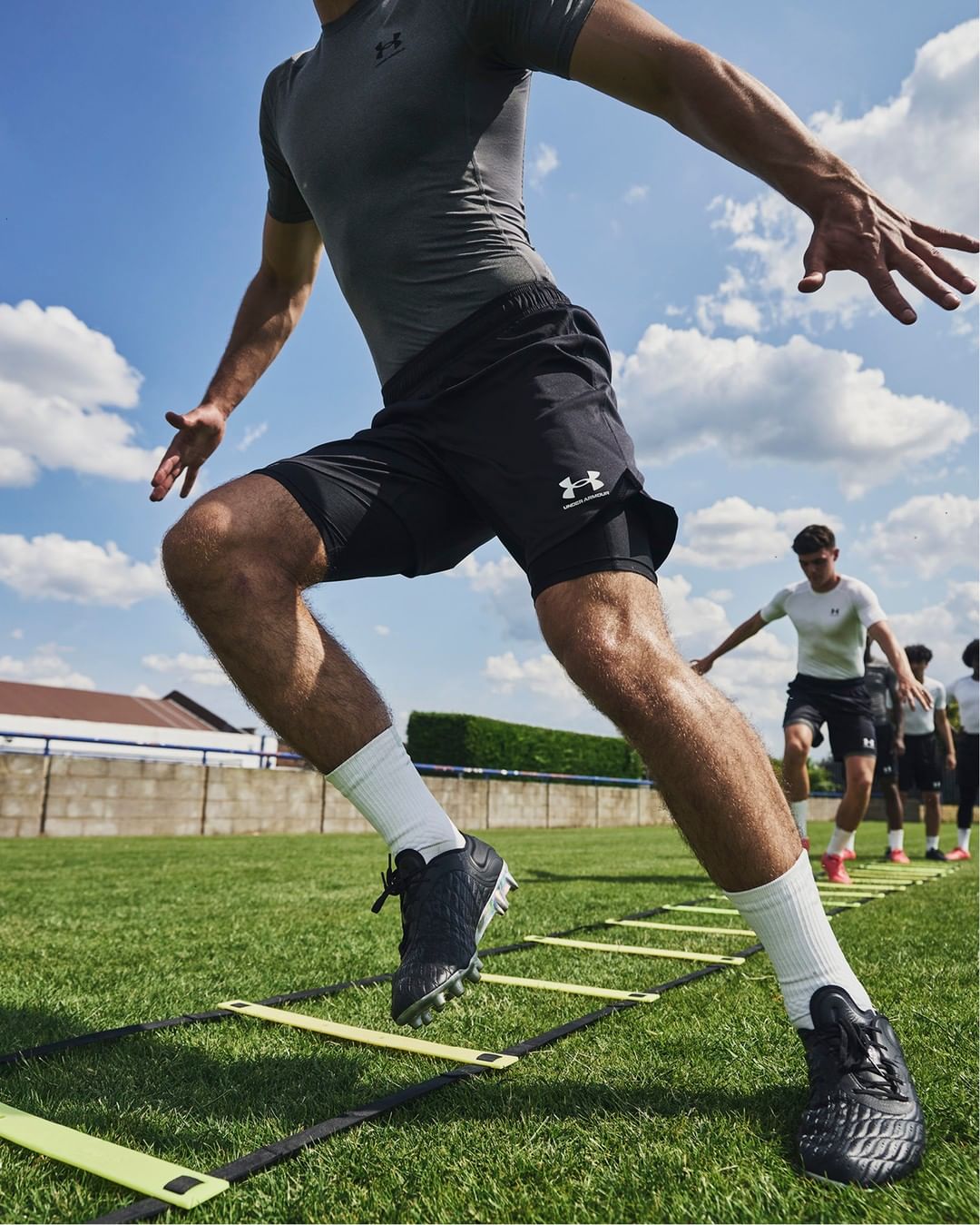 A man in athletic wear leaps over agility hurdles on a grass field under a partly cloudy sky, with other athletes in the background.
