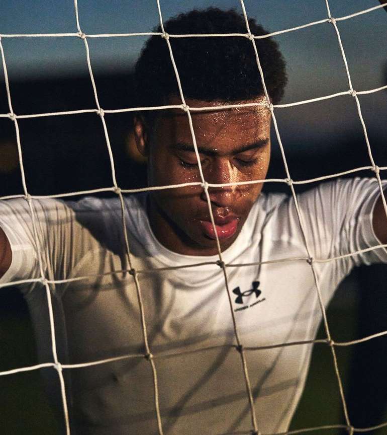 Close-up of a young adult male soccer player in white jersey leaning on a net with a focused expression and visible perspiration.