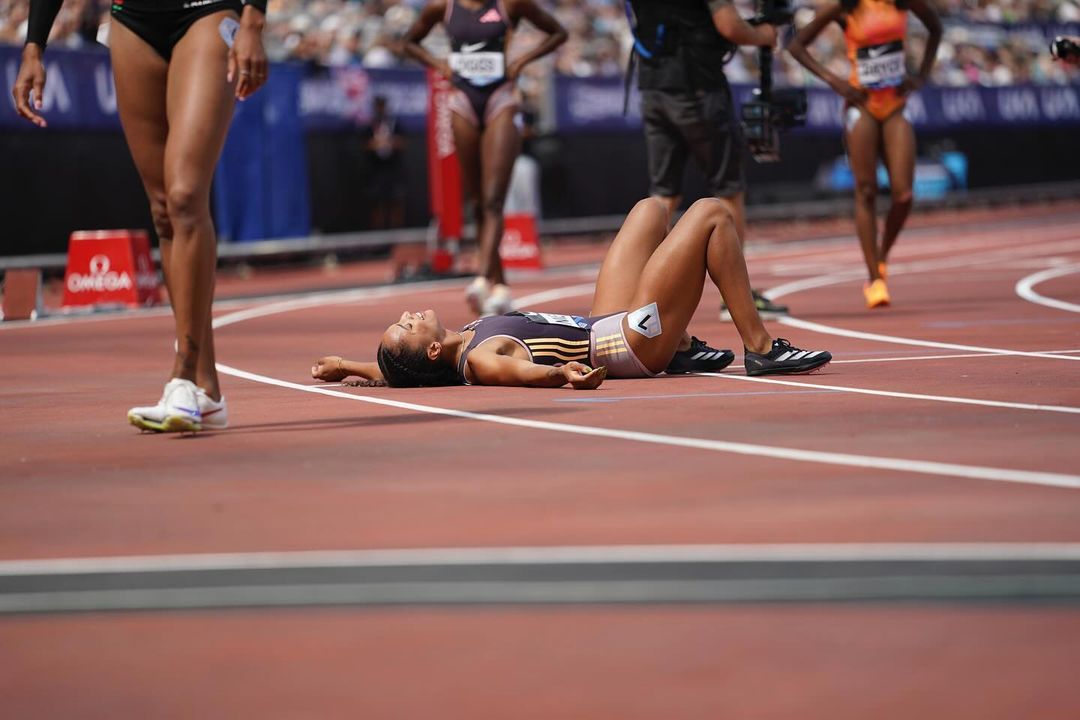 Female athlete lies on her back on a red running track, resting after a race; track competition with competitors in the background.