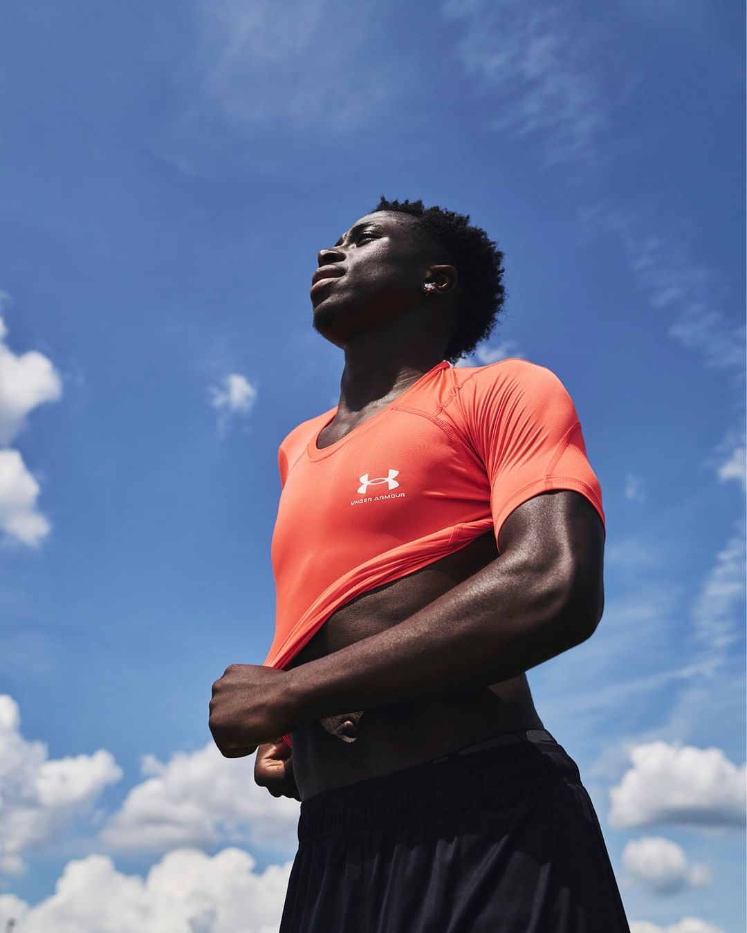 Athletic, dark-skinned man in orange shirt and black shorts looking up at the sky with white clouds. Sunny outdoor setting, low angle.