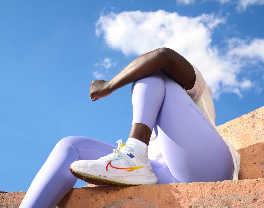 A running shoe and person are visible, with a bright blue sky and white clouds in the background, on a textured orange surface.