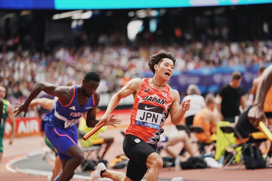 Athlete from Japan competing in a relay race, running with baton in hand on the track, crowd in background. Sports competition.