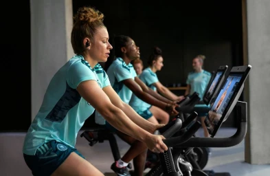 Group of female athletes in matching athletic wear riding stationary indoor bicycles during a workout session.