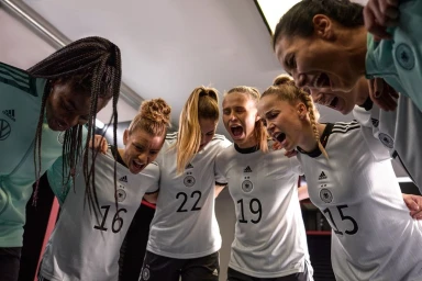 Group of six female soccer players in matching white jerseys huddled together in a locker room, shouting with passionate expressions.