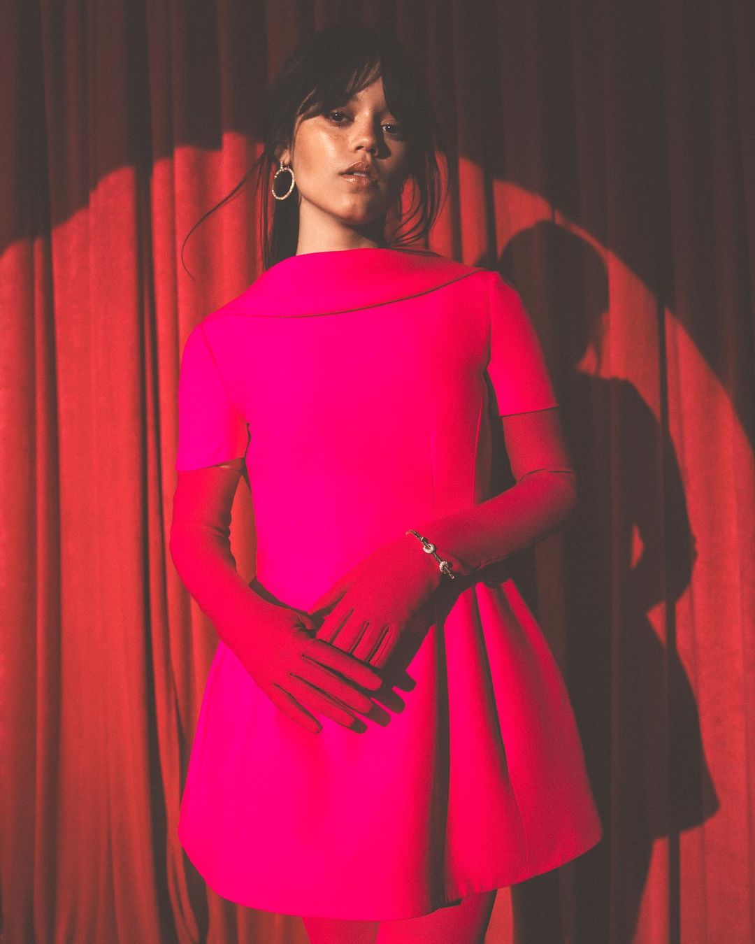 Portrait of a female in a bright pink dress with matching gloves against a red curtain backdrop, showcasing high fashion.