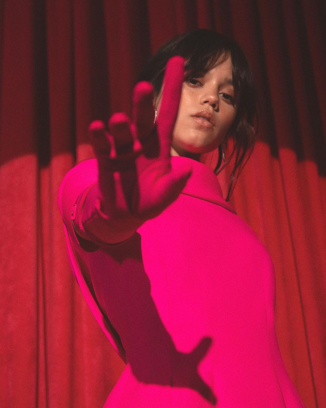Portrait of brunette adult female in hot pink jacket with arm extended toward the camera standing against red curtain in photo studio.