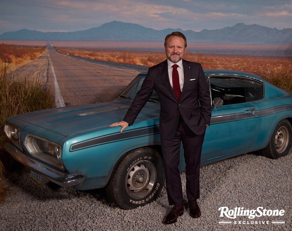 Man in suit leaning on vintage car against desert road backdrop, Rolling Stone logo in corner, creating a vintage aesthetic.