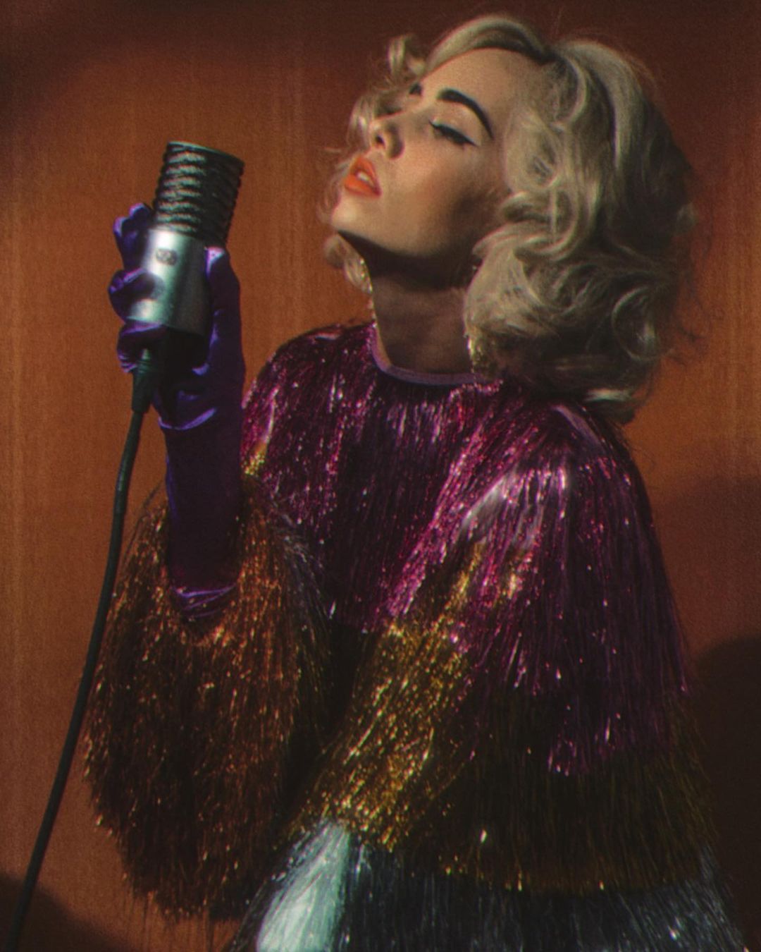 Woman with blonde curls sings into a vintage microphone, wearing a colorful tinsel dress and purple gloves against an orange background.
