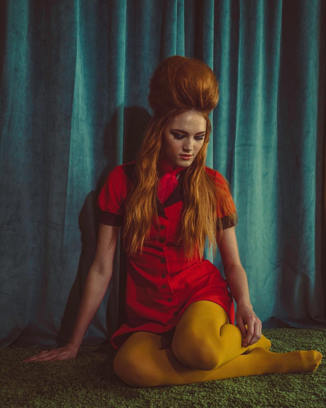 A redhead woman with a retro hairstyle wearing a red dress and yellow tights poses in front of a blue curtain in a studio shot.