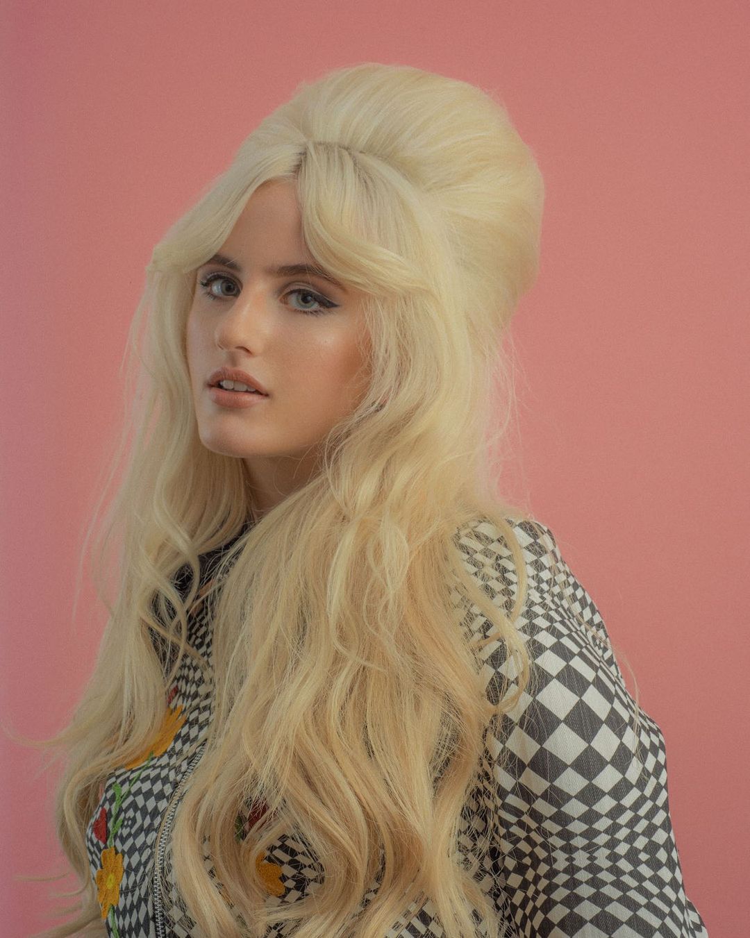 Portrait of a blond adult female with a bouffant hairstyle wearing a patterned jacket against a pink background in a photo studio.