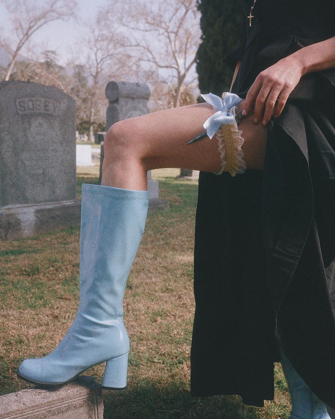 Fashionable woman in blue knee-high boot posing with garter in graveyard with tombstones and grass. Fashion or beauty editorial content.