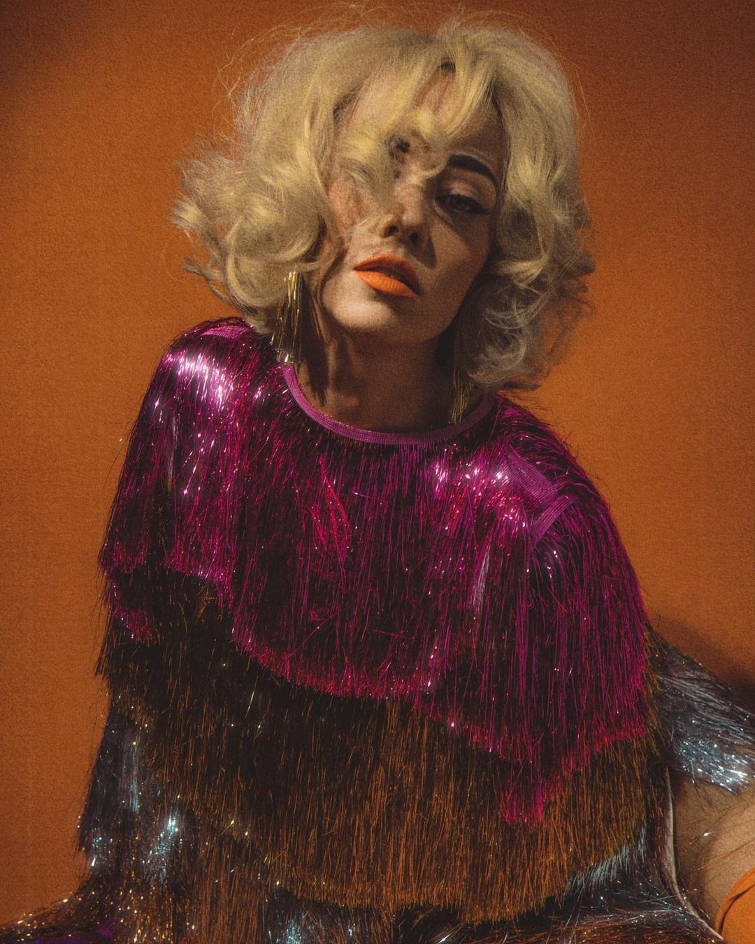 Portrait of a blond adult female wearing a sparkling multi-colored top against an orange background in a studio setting.