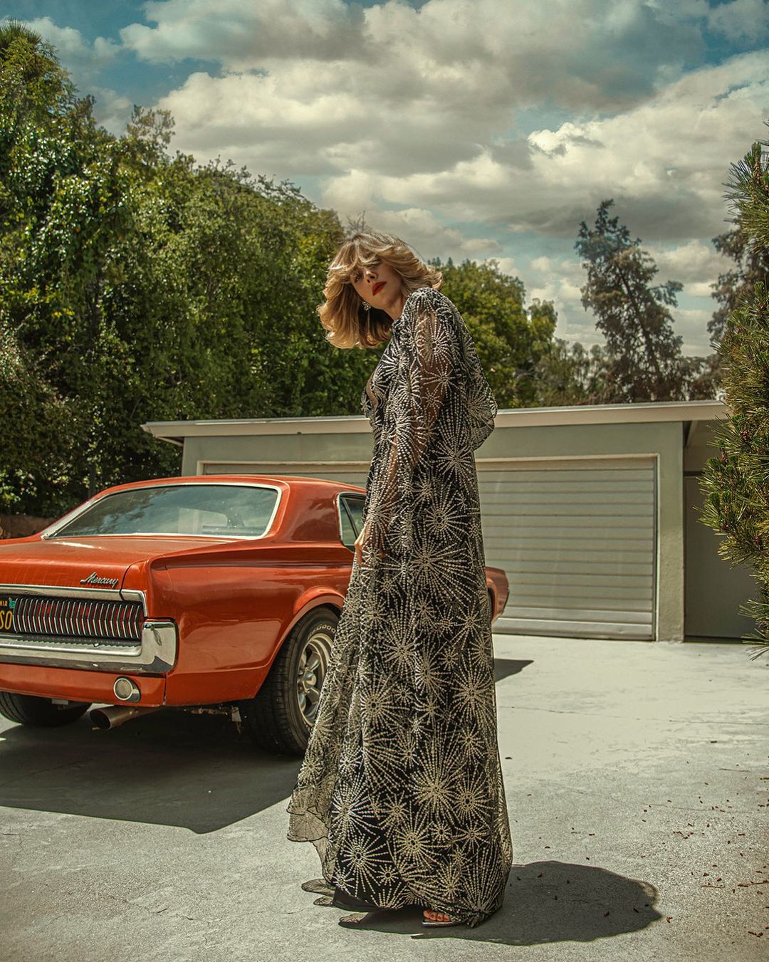Adult woman in flowing sheer dress with car on driveway. She has blonde hair and is wearing red lipstick. Trees and sky background.