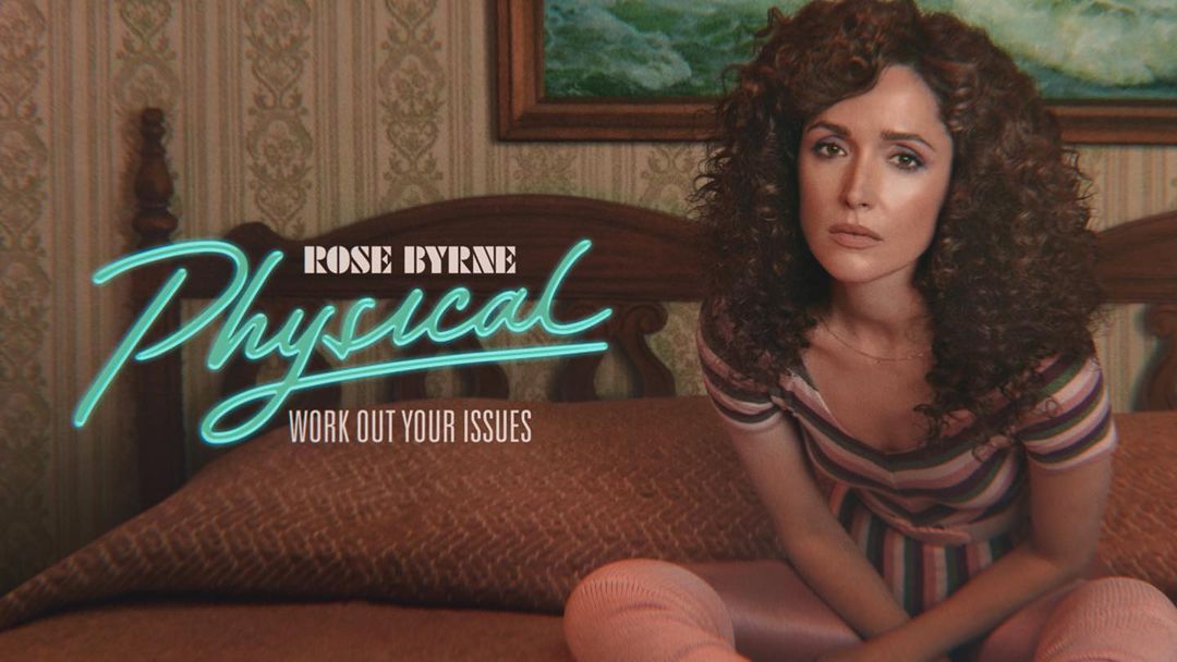 Rose Byrne poses indoors for the 'Physical' TV series. She wears a striped shirt and pink leg warmers, sitting on a patterned bed.