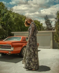 Blonde woman wearing a long patterned dress standing outdoors beside a vintage orange muscle car.