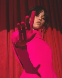 Jenna Ortega in a vivid pink dress with a gloved hand extended forward, set against a pleated red curtain background.
