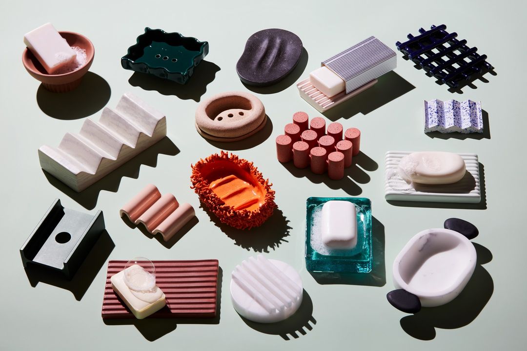 Assortment of colorful soap dishes in various shapes and materials are arranged on a soft green surface in studio lighting.