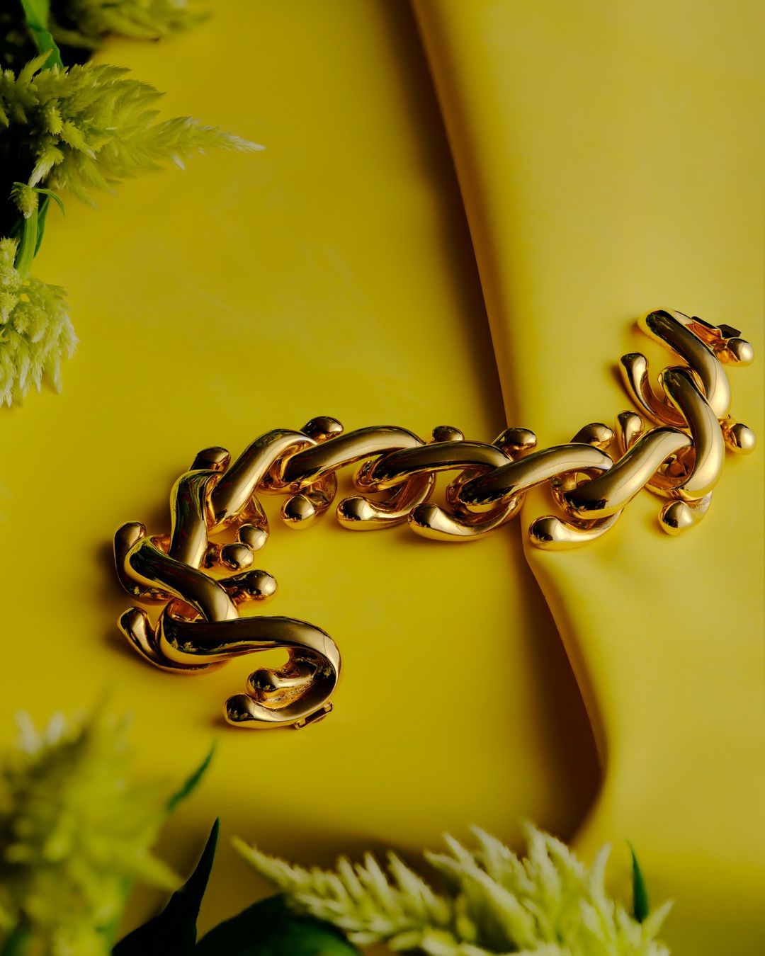 A bold gold chain bracelet is staged on a luminous yellow backdrop with soft green floral accents, highlighting jewelry design.
