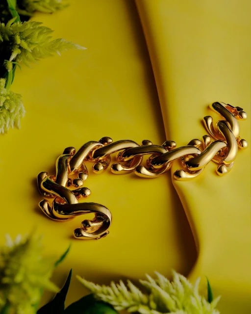 A bold gold chain bracelet is staged on a luminous yellow backdrop with soft green floral accents, highlighting jewelry design.