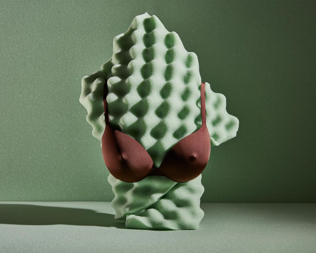 Still life of a dark-red bra draped on a light-green textured foam sculpture against a matching light-green backdrop.