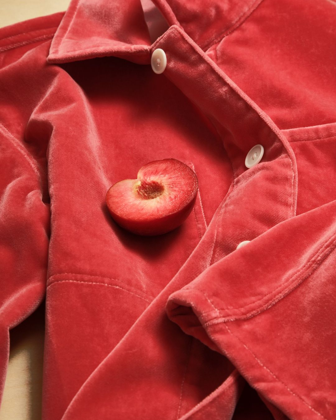 Close-up of a sliced plum placed on a folded, red velvet jacket with white buttons, creating a luxurious, textured, and minimalist composition.