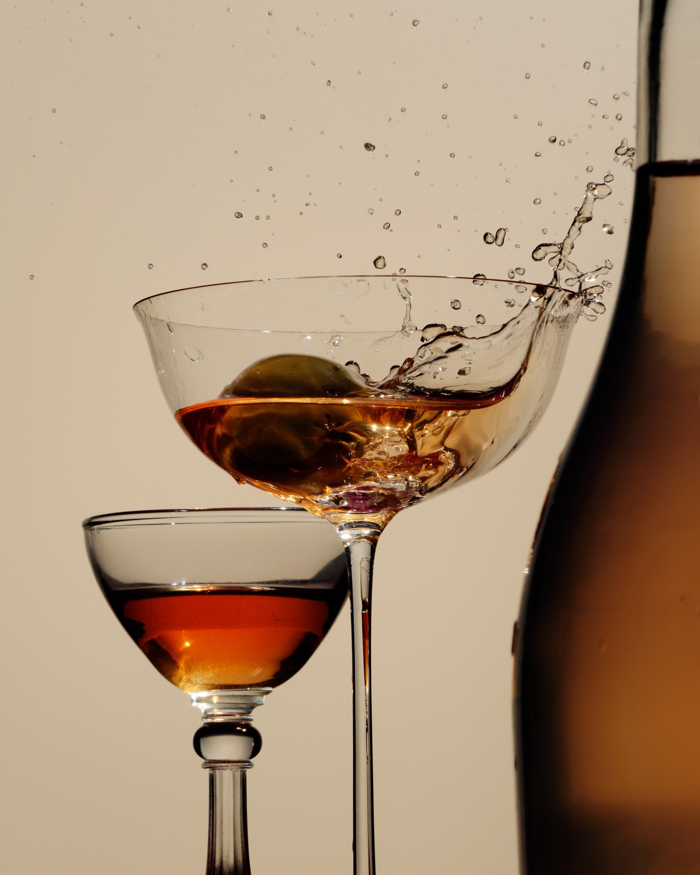 Close-up studio shot of a stemmed glass of amber liquid with an olive splashing, alongside a second glass and a bottle.