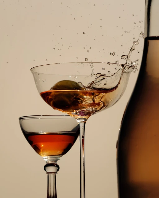 Close-up studio shot of a stemmed glass of amber liquid with an olive splashing, alongside a second glass and a bottle.