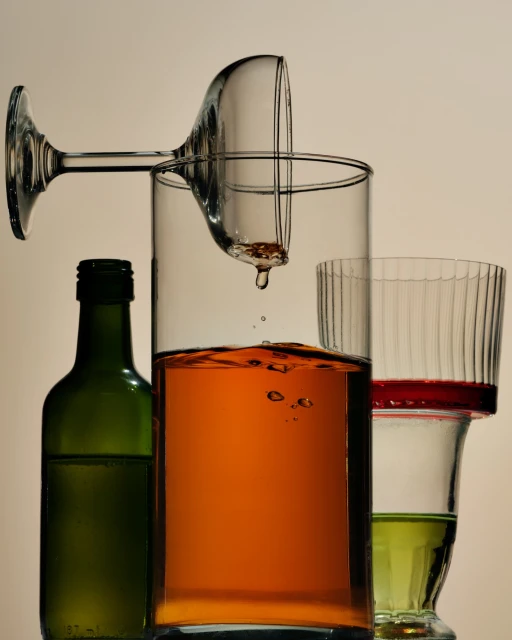 A still life composition featuring various glassware, including a tipped martini glass, a tall cylindrical glass, and a green bottle.