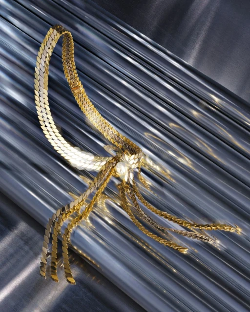 A gold metallic-linked necklace cascades over a wavy silver surface, reflecting light in a studio fashion shot.