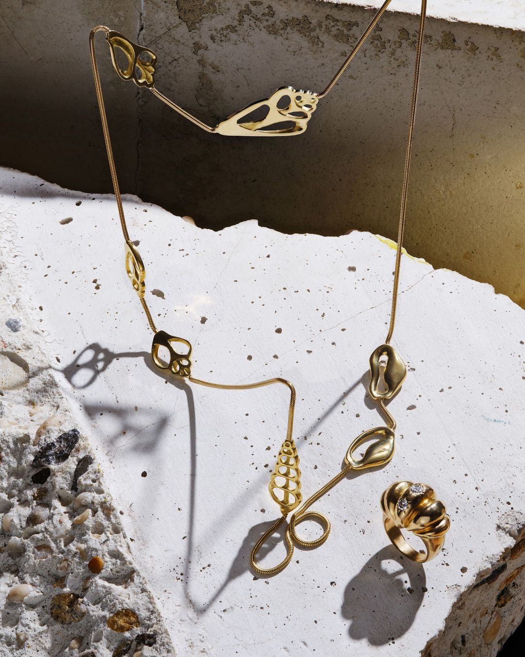 Gold necklace with abstract charms paired with a textured gold ring on a fractured concrete surface under studio lighting. Fashion jewelry.