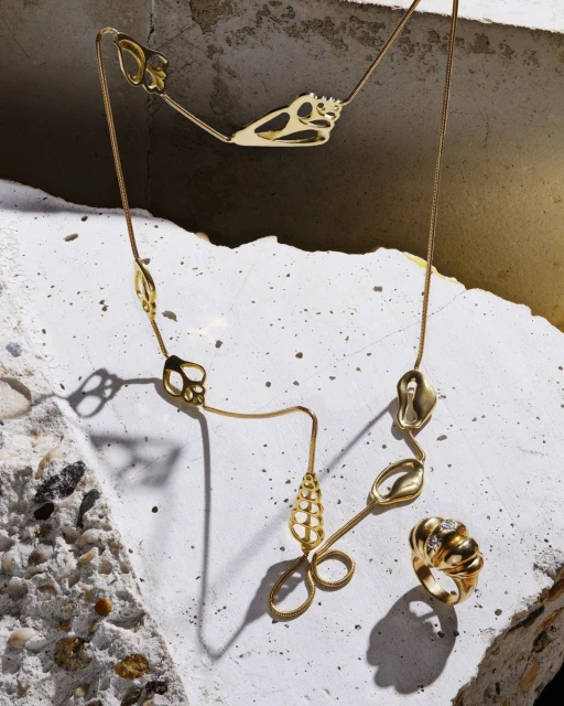 Gold necklace with abstract charms paired with a textured gold ring on a fractured concrete surface under studio lighting. Fashion jewelry.