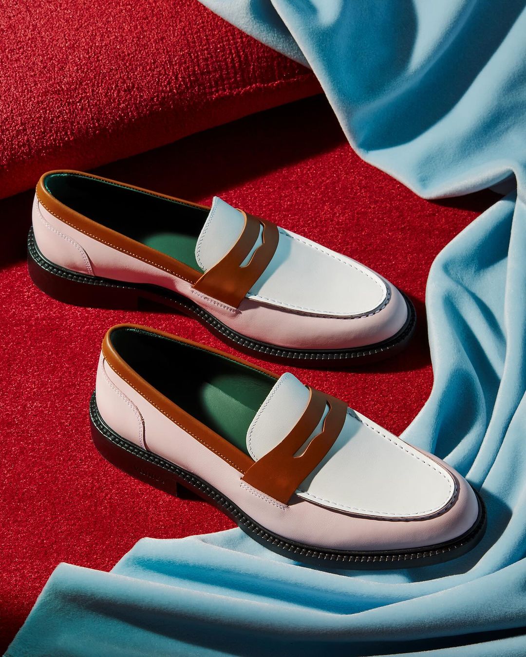 Two-tone pink and white leather penny loafer shoes sit on a red velvet surface near a draped light blue silk fabric.