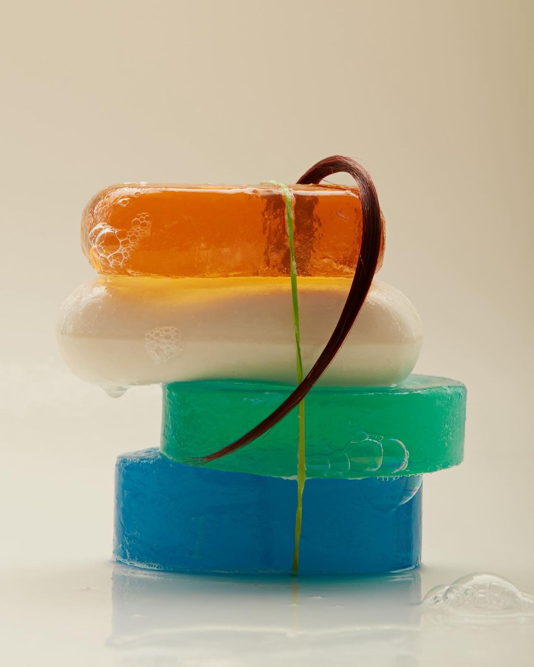 Stack of transparent soaps with orange, white, green, and blue colors and a curved brown stem, arranged on a reflective surface.