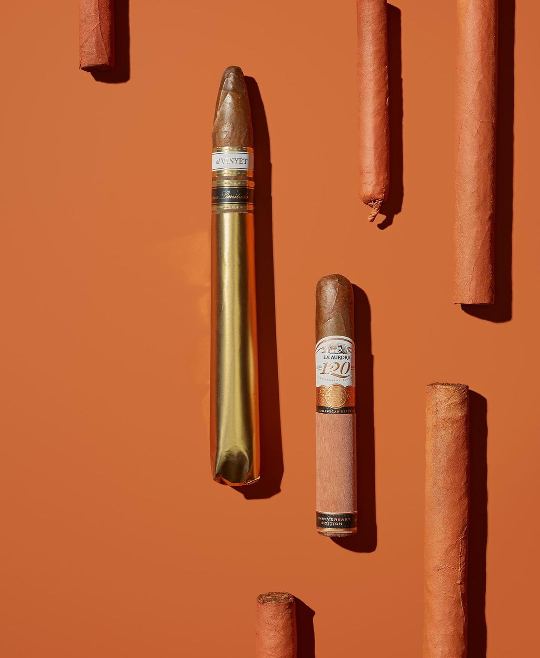 Close-up studio shot of several cigars varying in size and wrapper, set against a flat orange backdrop with soft, diffused lighting.