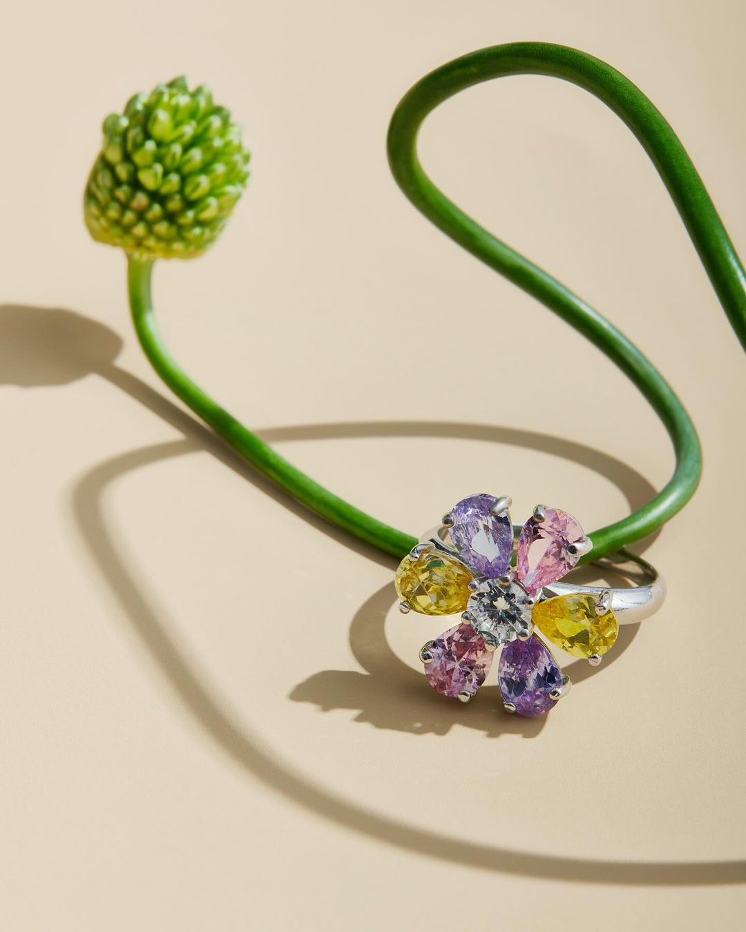 Close-up of a flower ring made of yellow, pink, and purple gemstones next to a curved green flower stem and bud on a cream background.